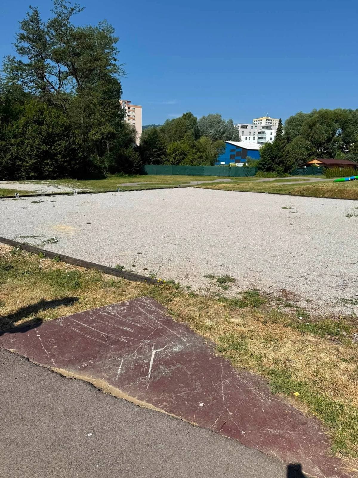 Prázdna spevnená plocha so zvyškami asfaltového povrchu, v pozadí zeleň a panelové domy